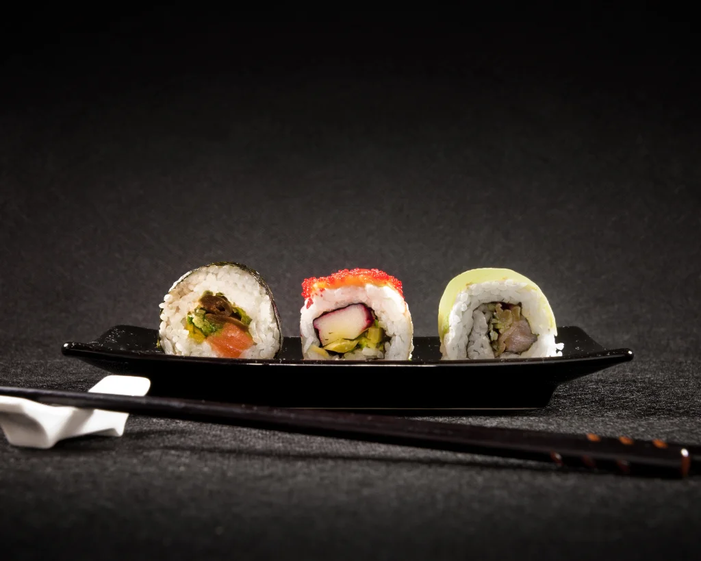 Ein Maki Sushi & zwei Uramaki liegen luxuriös angerichtet auf einem schwarzen, länglichen Teller. Davor schwarze Essstäbchen.