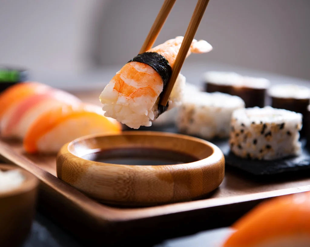 Sushi-Set auf einer Holzplatte, im Vordergrund Bambusessstäbchen, ein Shrimp-Nigiri wird in ein Schälchen Sojasauce getaucht.